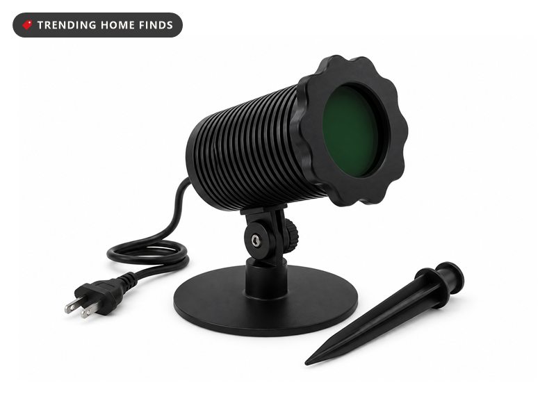 Black outdoor light projector with a ribbed barrel, a scalloped front bezel and a green lens, on a ground stake beside its round base.