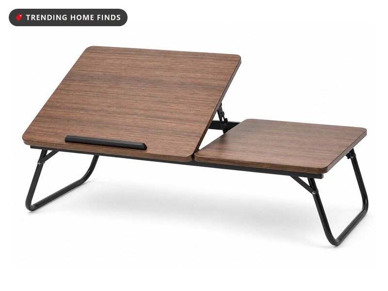 Black folding lap desk on a bed with a laptop on its tilted left panel and a phone and cup slot on the flat right side.