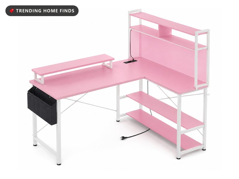 Pink L-shaped gaming desk on a white metal frame, with a raised monitor shelf, a two-tier hutch carrying a power strip, a hanging fabric side pocket and two lower shelves