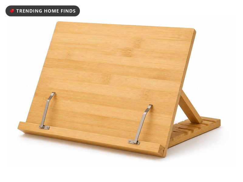 A pale bamboo book stand holding an open book upright, with a notched prop arm behind and two metal page clips on the ledge.