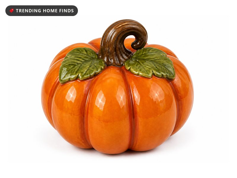 A glossy orange ceramic pumpkin with rounded ribs, two green leaves and a curled brown stem.