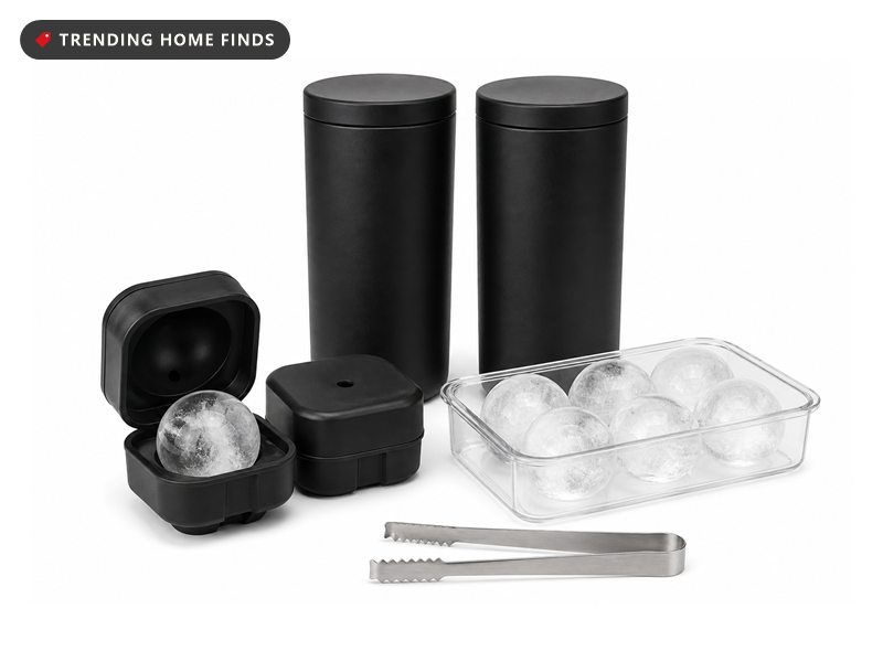 Clear ice ball maker moulds with finished ice spheres in a storage tray, steel tongs, and a whiskey glass holding a clear ice ball