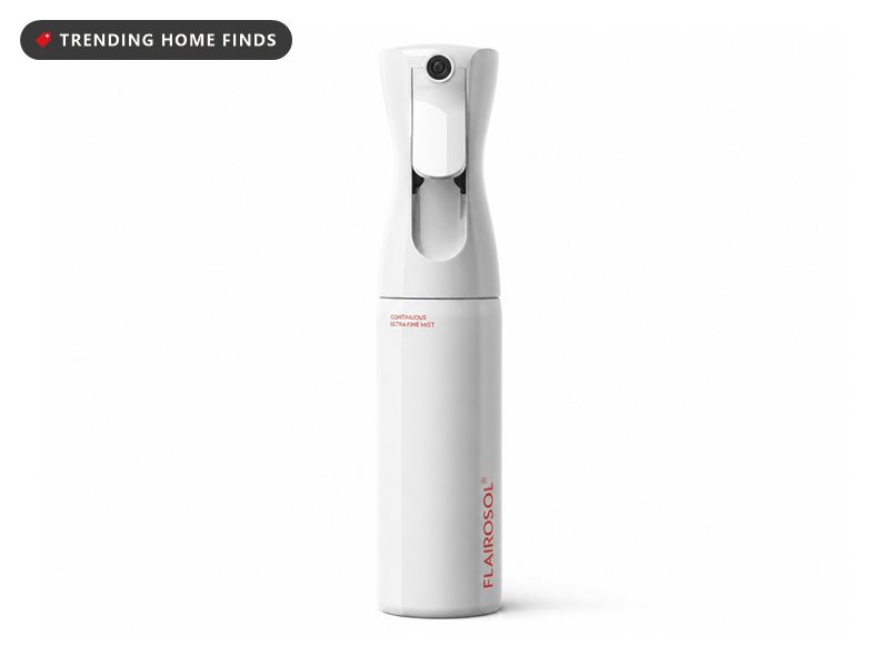 A white continuous mist spray bottle held up to long damp brown hair in a warm bathroom, the spray crossing the lengths.