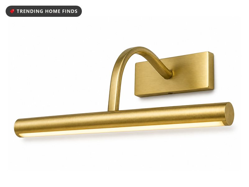 A slim brushed gold picture light bar on a curved arm bracket, switched on with a warm glow along its lens.
