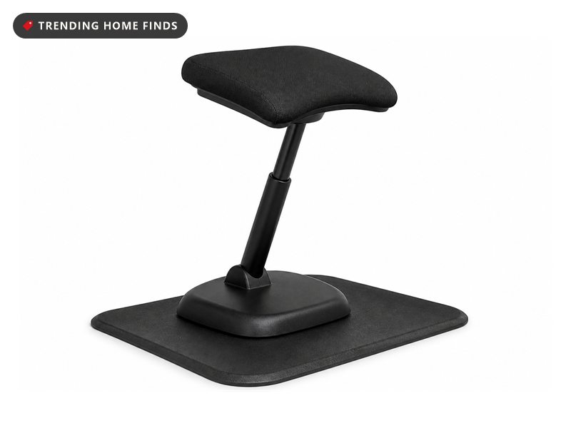 A black leaning perch stool with a curved seat tilted at a raised standing desk by a window in golden hour light, its post angled forward.