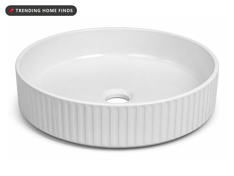 A white fluted ceramic vessel sink on a wood powder room vanity under an arched brass faucet, late night light raking across the ribs.