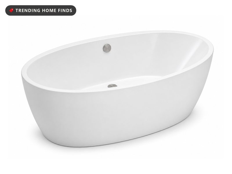 A glossy white oval freestanding tub against a fluted plaster wall in bright midday light, eucalyptus in a jug and a low wooden stool.