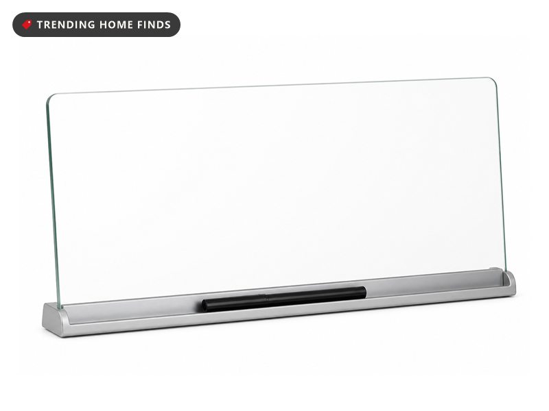 A slim glass memo board angled between a keyboard and a monitor on a pale desk in midday light, a marker resting in its storage channel.