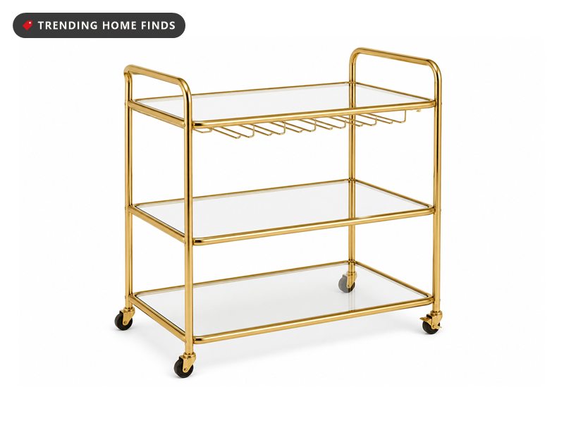 A gold three tier bar cart with clear glass shelves and a rack for hanging glasses.