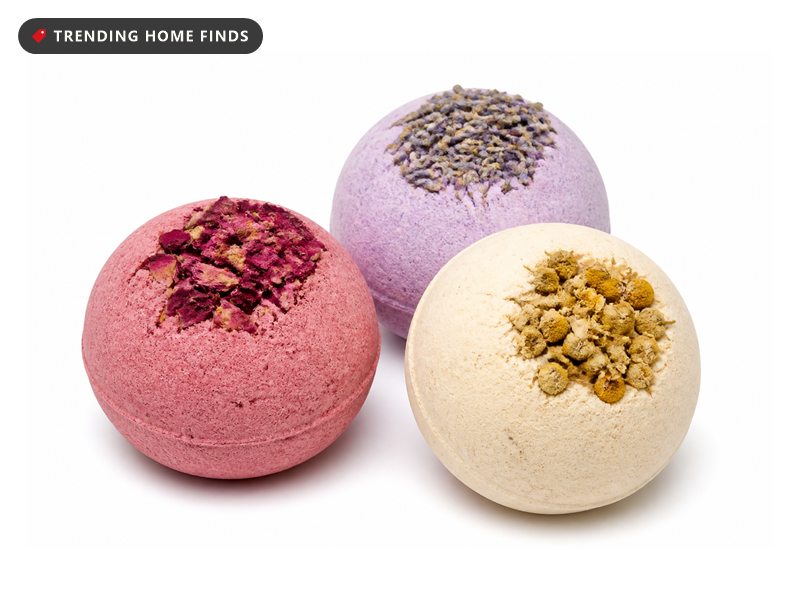 Three flower topped bath bombs on a folded towel beside a filled bathtub, colour blooming through the water in warm golden hour light.