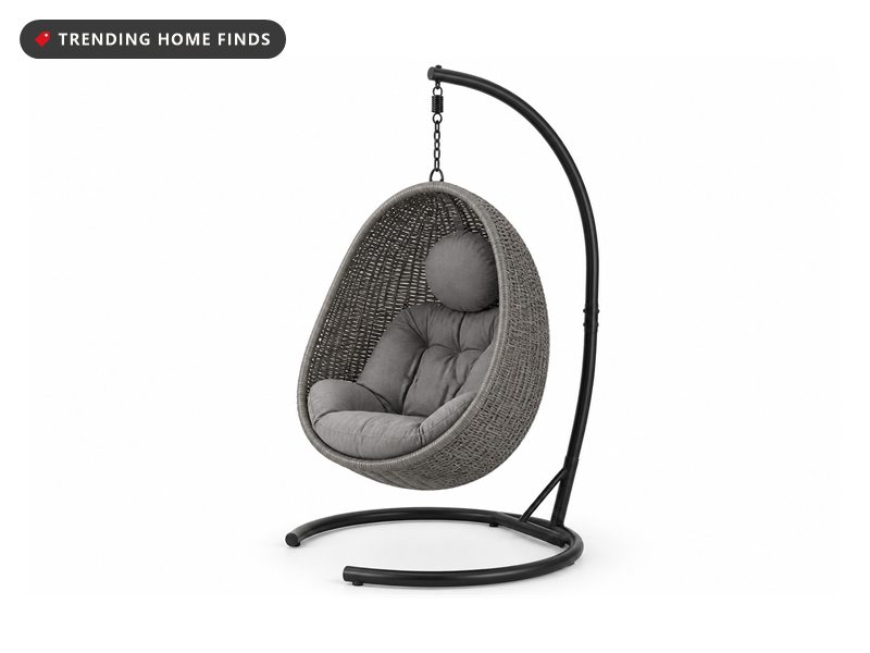 A grey wicker hanging egg chair on a black steel stand under a covered patio in soft light after rain, jute rug and olive tree beside it.