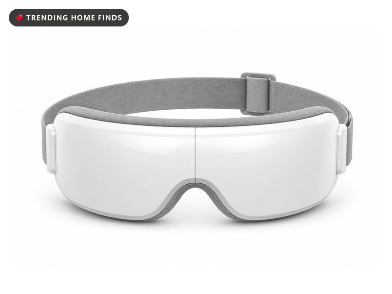 White heated eye massager mask with a curved padded interior, shown folded flat beside its case