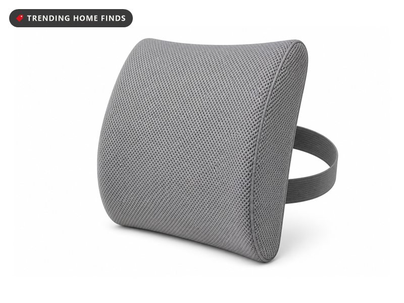 A grey contoured memory foam cushion strapped to the back of a dark office chair beside a desk, flat overcast daylight from the window behind.