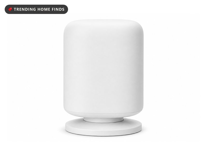 A small white presence sensor on a living room bookshelf beside a lit lamp in bright midday light, part of a smart lighting setup.