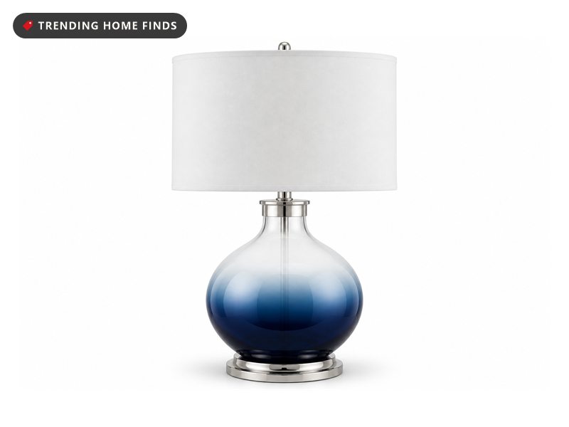 A blue ombre glass table lamp with a white drum shade lit on a console table against a dark charcoal wall at golden hour.