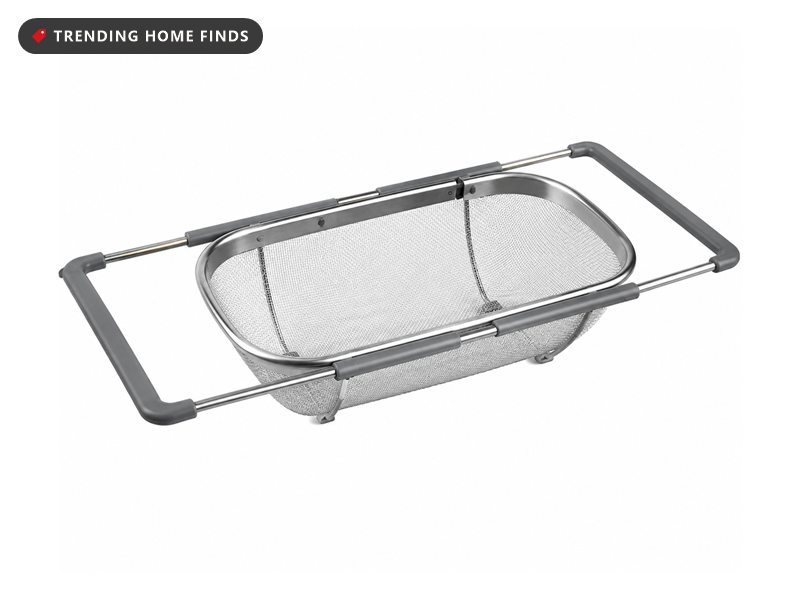 Stainless steel mesh colander with grey extendable side arms, resting across the width of a sink