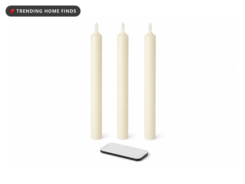 Three ivory flameless taper candles flickering in mismatched brass holders on a painted brick mantel by a rain-streaked window.