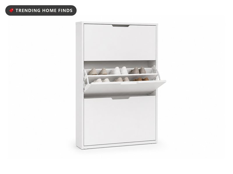 White shoe cabinet in an entryway with all three flip drawers tipped open showing rows of shoes, a clock and plant on top.