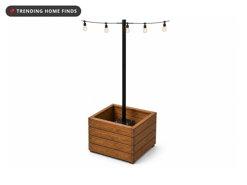 A tall black steel light pole rising from a square wood planter box of flowers on a wet patio, bistro lights swagging away from the top.