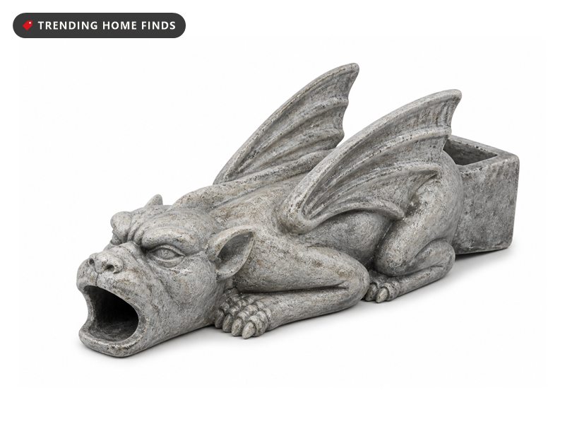 A gray stone look resin gargoyle downspout extension with folded wings and an open mouth, sitting on river rock.