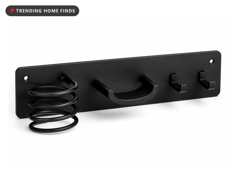 A matte black wall bar with a spiral coil, a center cradle and two hooks holding a dryer, a wand and a flat iron.